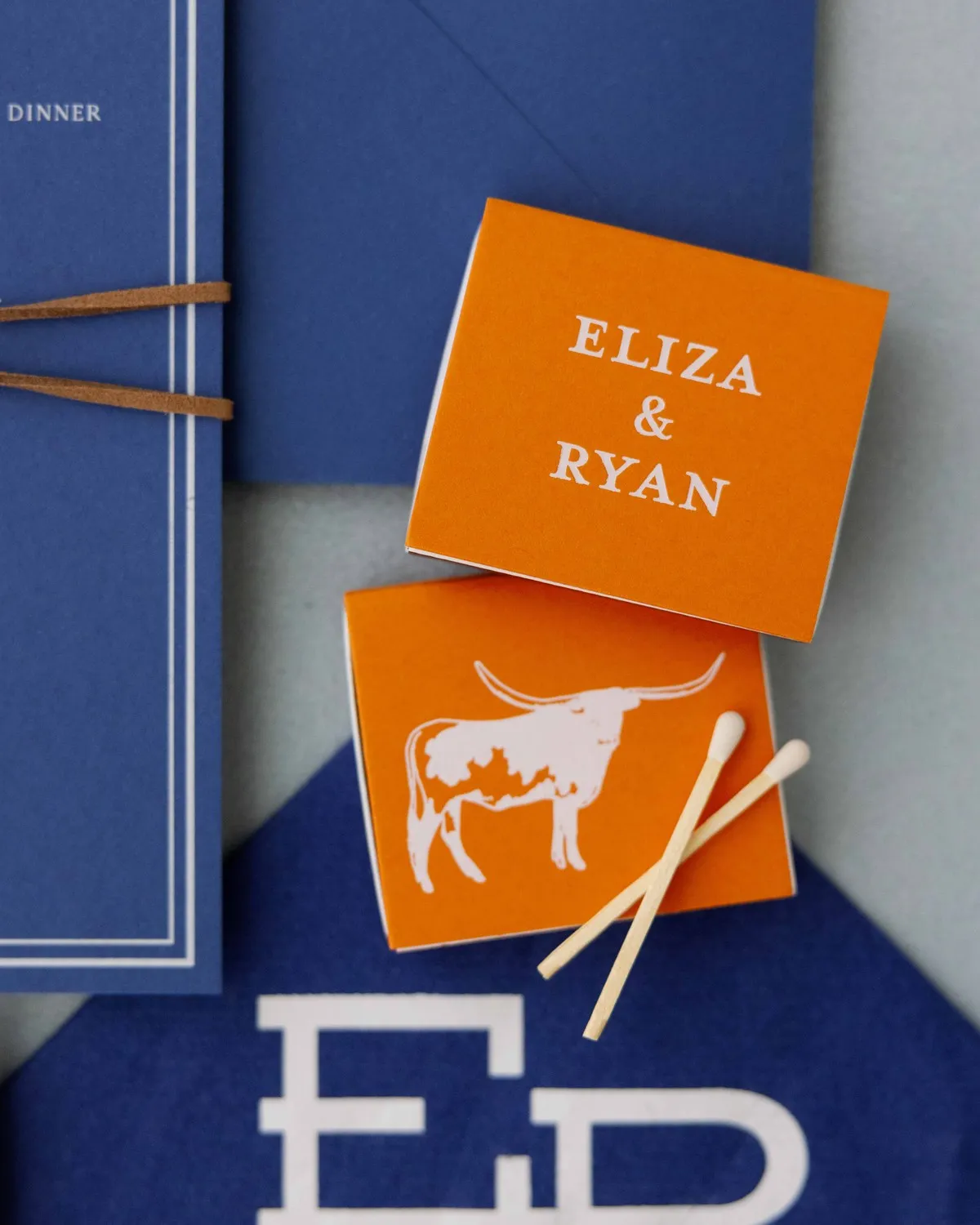 Orange longhorn matchbooks resting on a navy cocktail napkin with the Eliza monogram